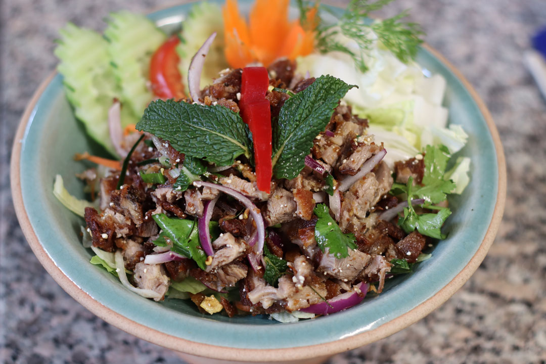 A close-up of a bowl filled with a salad featuring chopped meat, fresh herbs, and vegetables, garnished with mint leaves and red pepper slices.